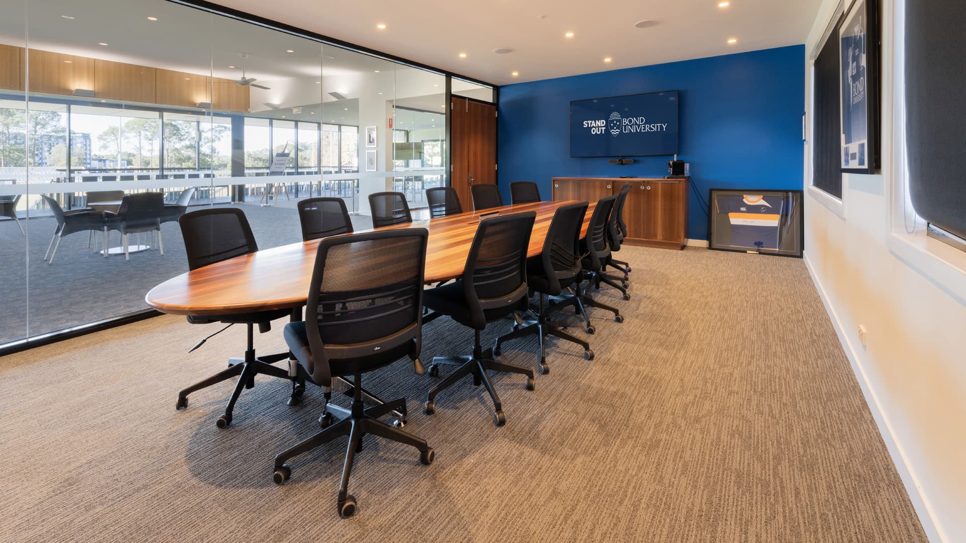 A wide angle view of a meeting room at Bond University with a long table, swivel chairs and a TV screen.