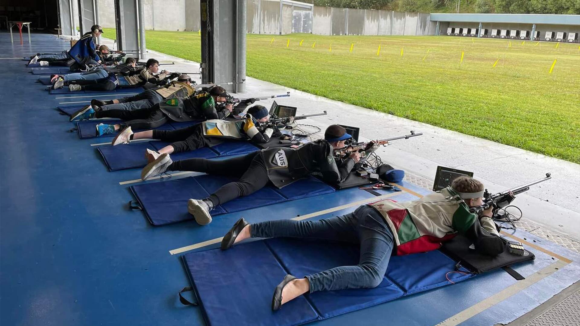 Ten people laying on the floor in a row shooting at targets across a field at Brisbane International Shooting Centre.