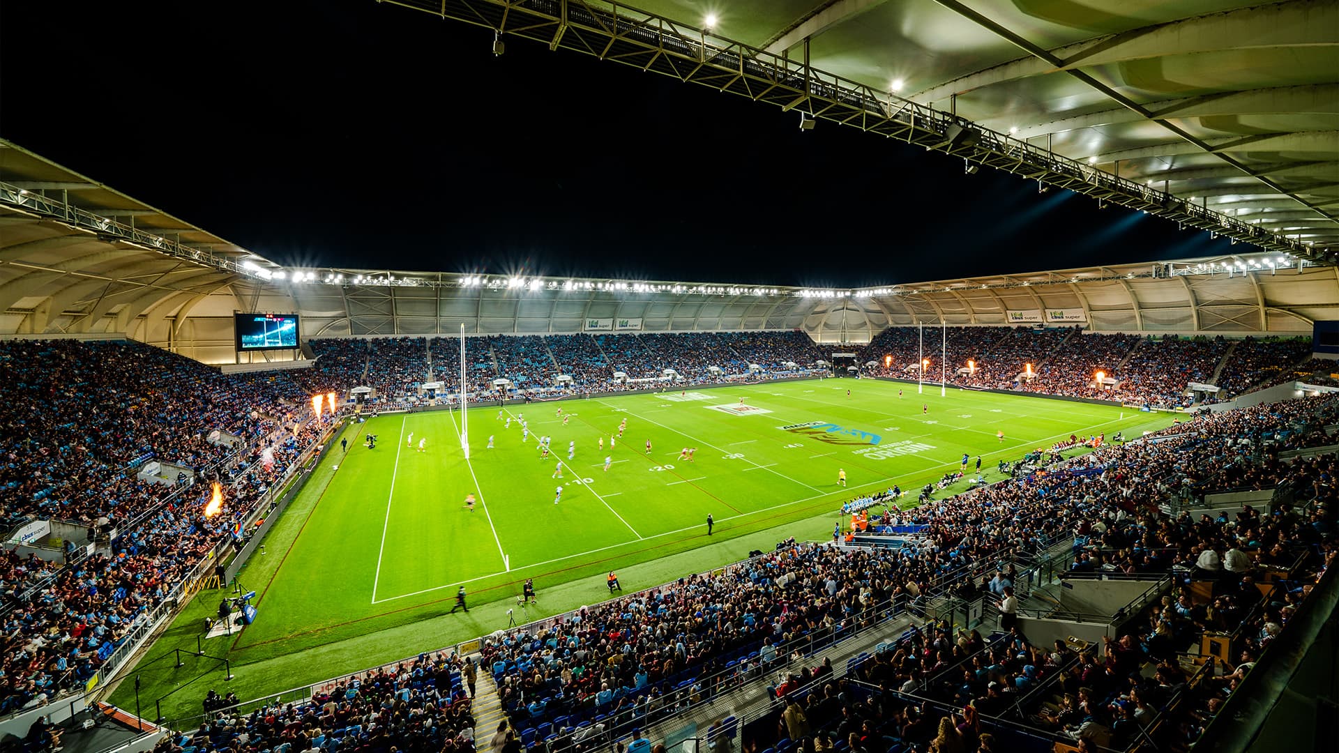 A wide angle view of CBUS Stadium during State of Origin with full spectator stands and players gathered at one end of the field.