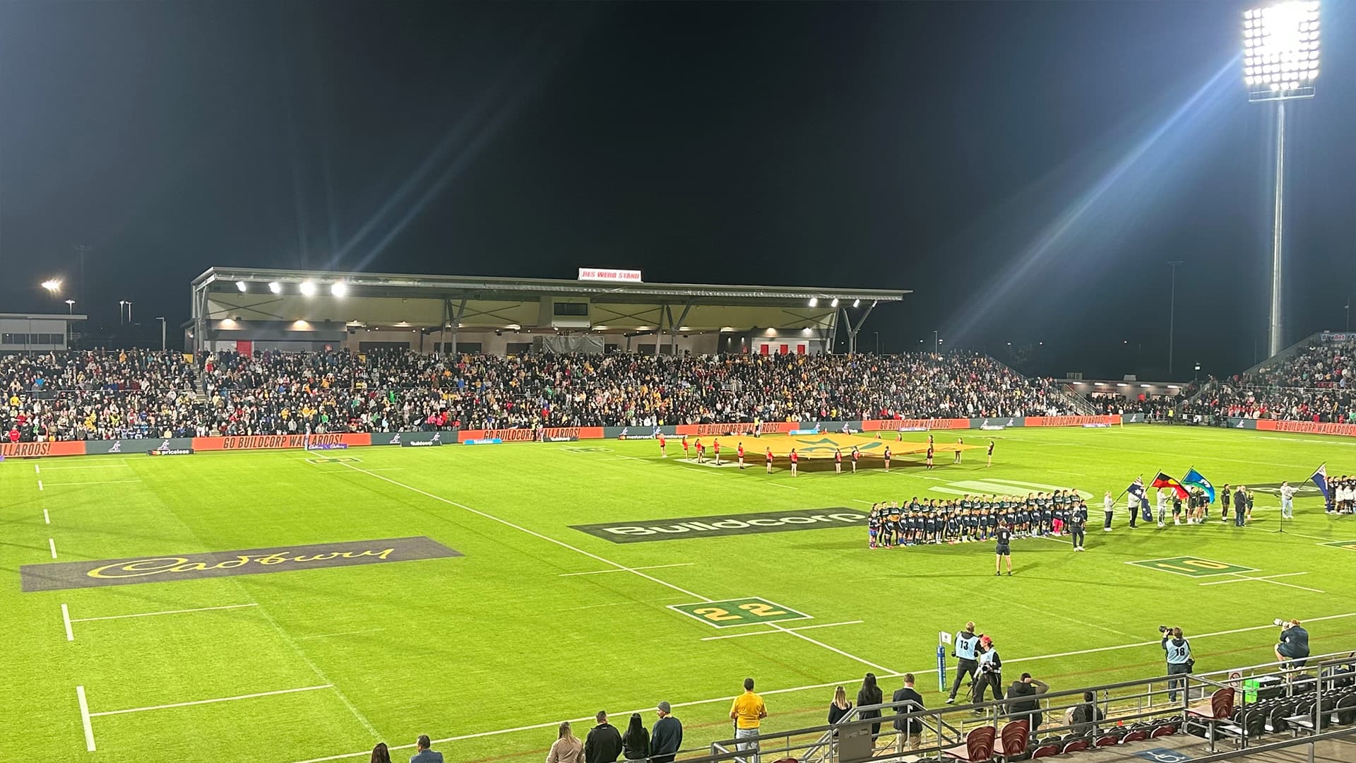 Kayo Stadium during a night game with fans and players standing for the national anthem. 