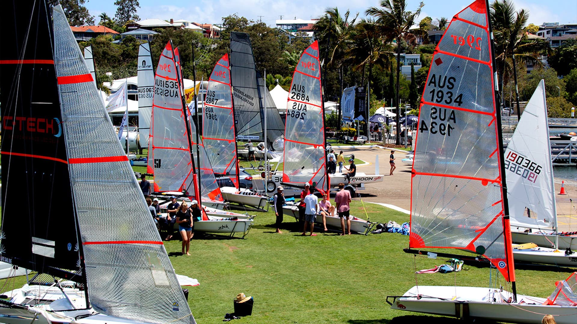 Eleven yachts on the grass next to the dock at Royal Queensland Yacht Squadron with people sitting on the boats.