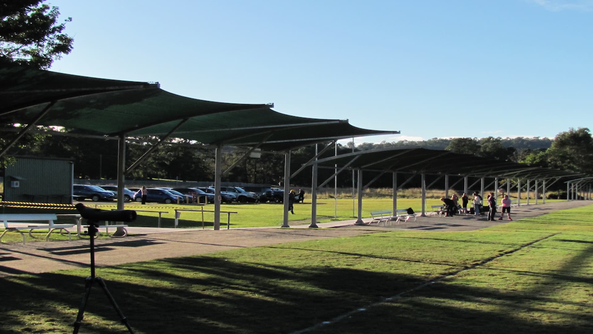 A group of people standing under the outdoor undercover area in front of the archery field. 