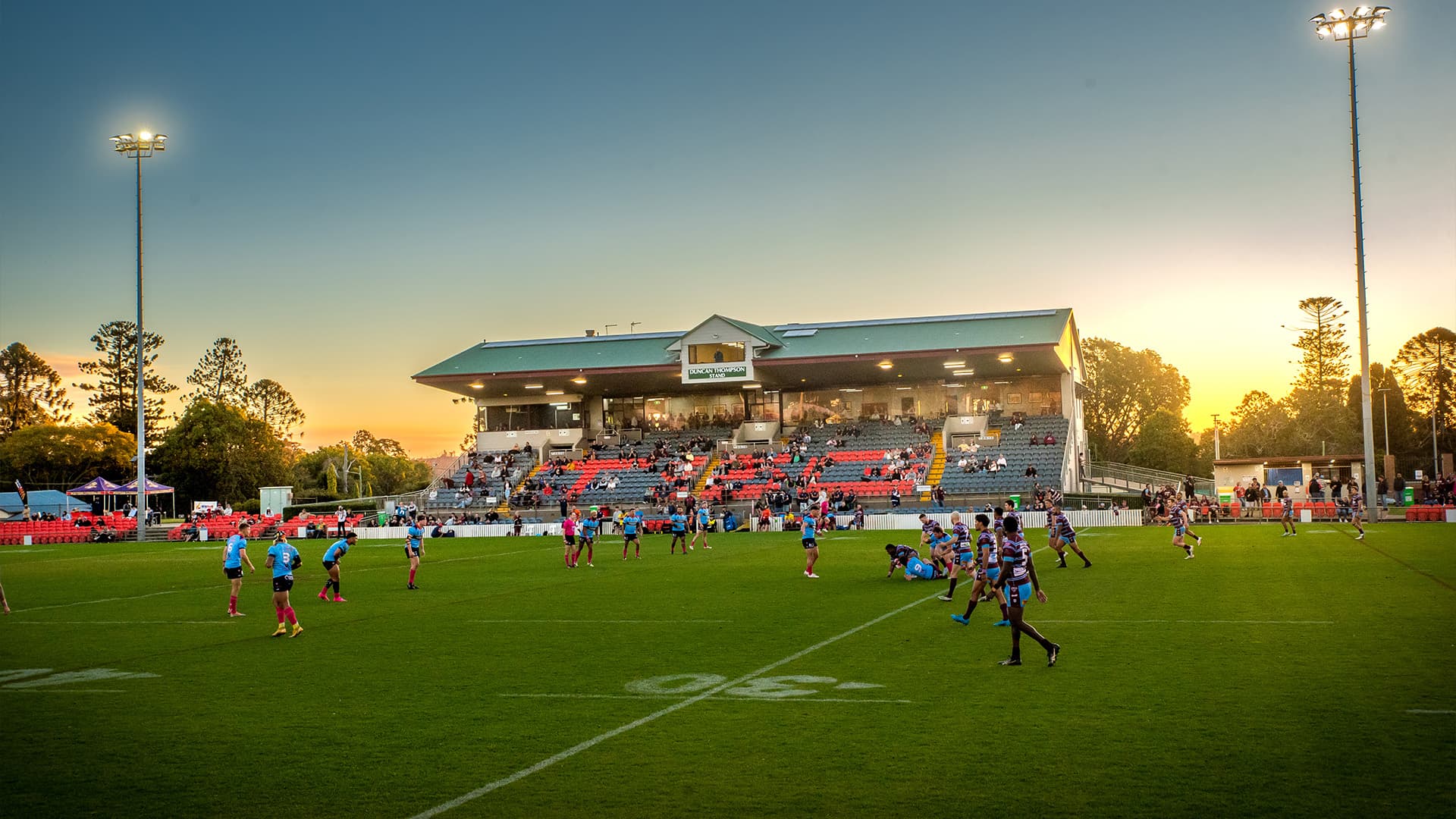 A wide angle view of a Rugby game at Toowoomba Sports Ground at sunset with players spread across the field and spectators in the stands.