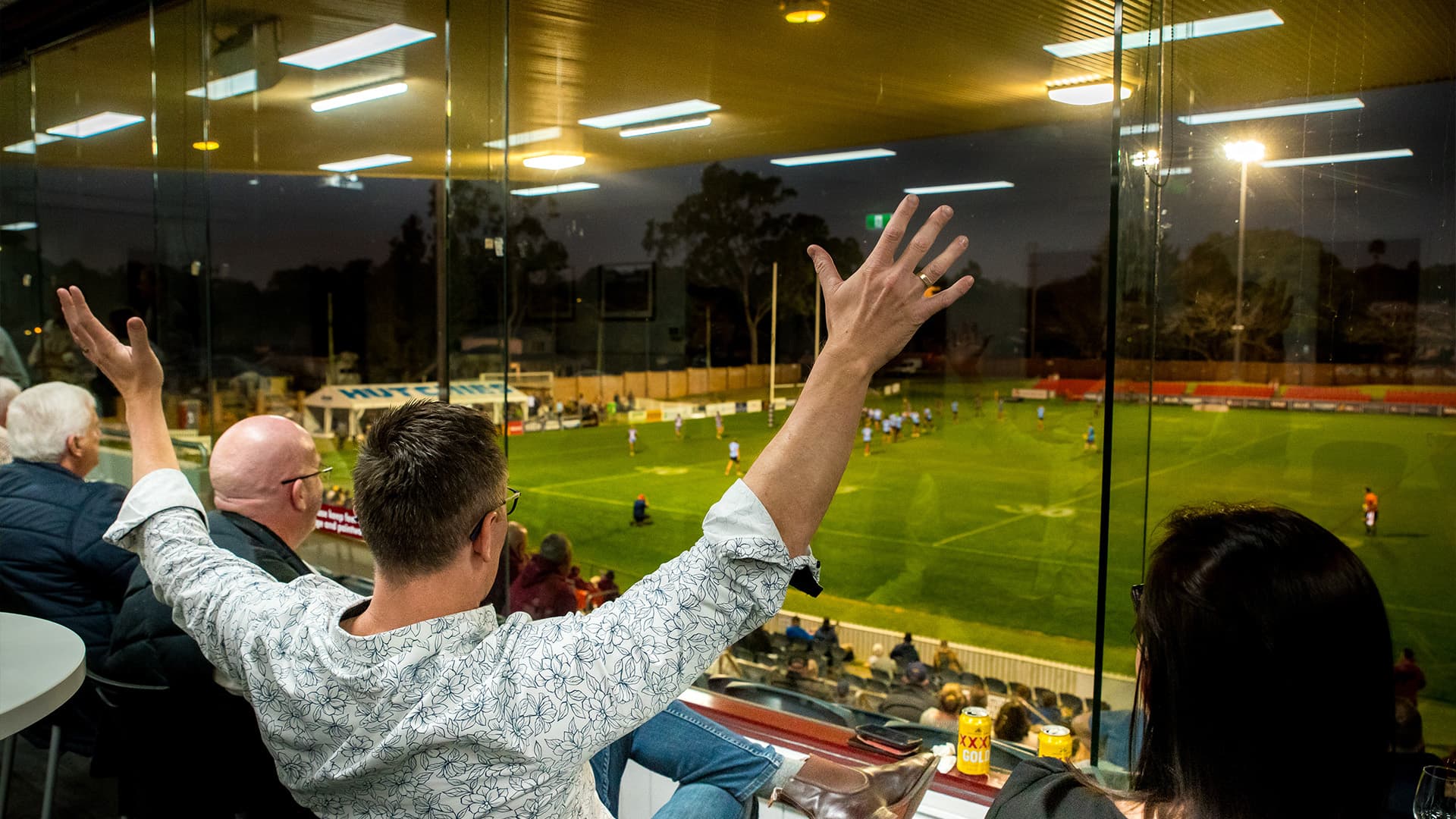 Man raising his hands in the air while watching the Rugby game from an indoor spectators box at the Toowoomba Sports Ground Field.